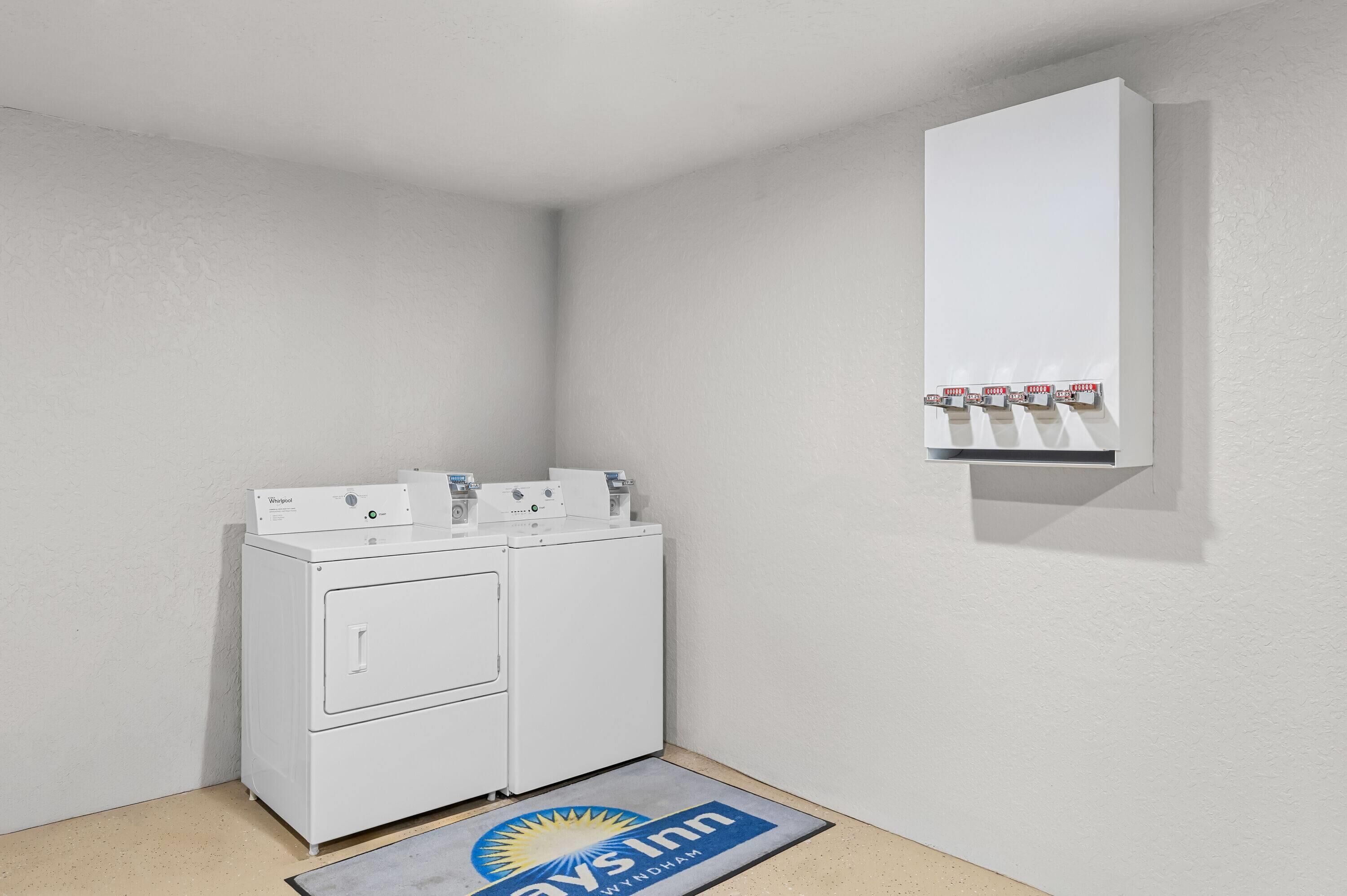 Une buanderie équipée d’un lave-linge et d’un sèche-linge à chargement frontal, d’une petite étagère avec des articles de toilette et d’un tapis orné du logo de l’hôtel.