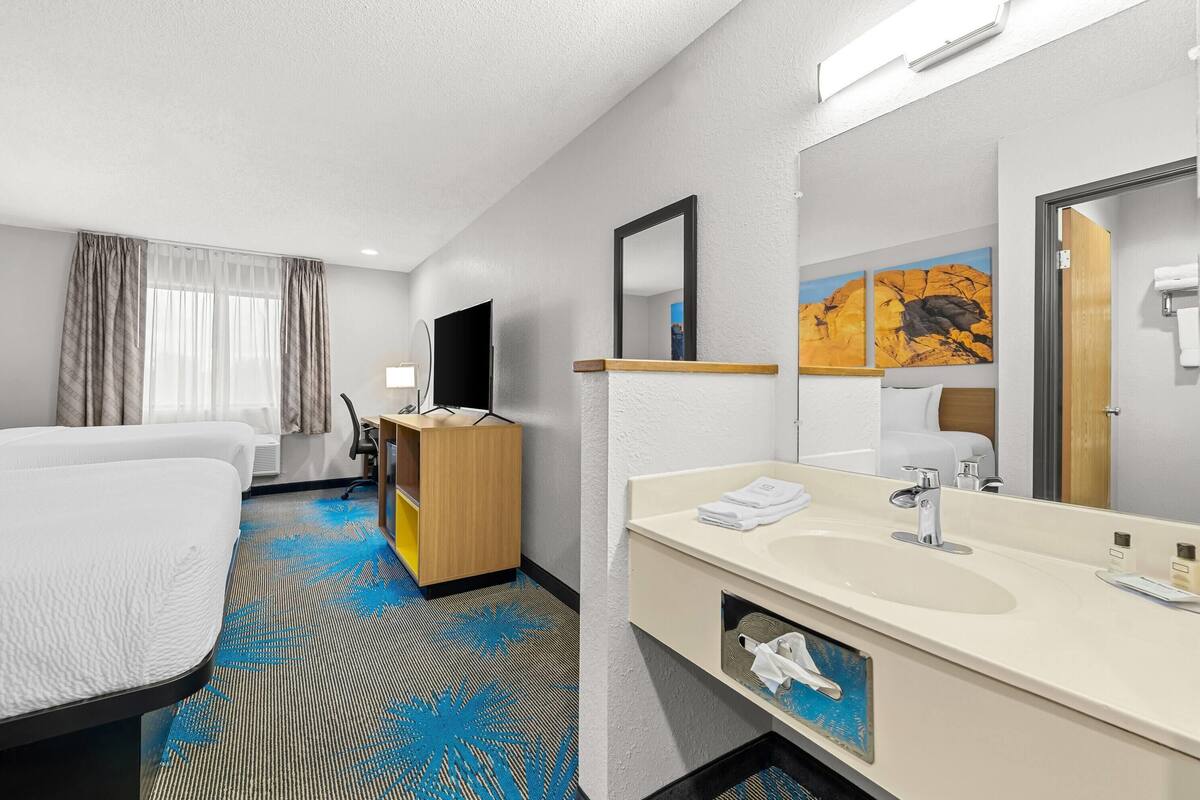A hotel room with a bed, a desk, a large mirror, a sink, and a window with curtains.
