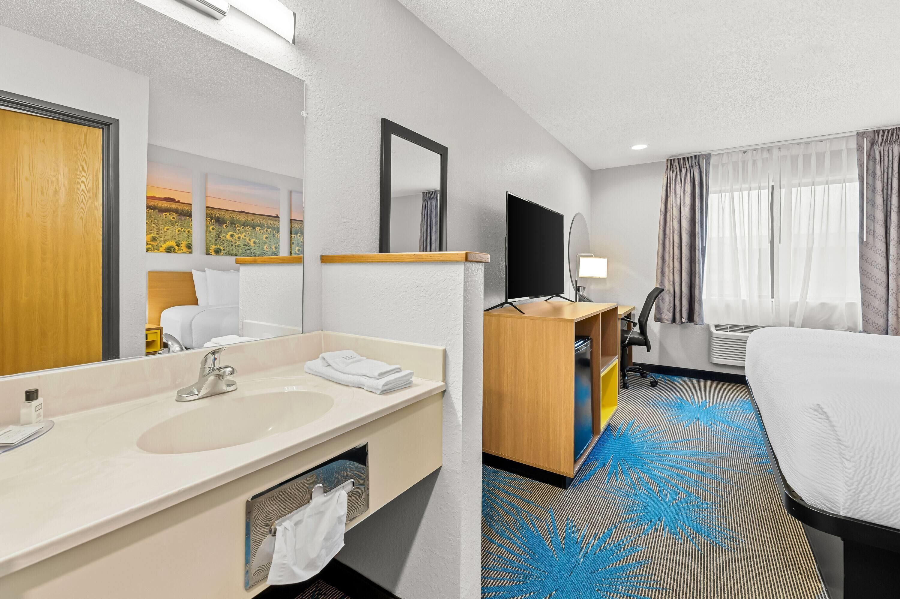 Habitación de hotel con una cama grande, baño con lavamanos y espejo, escritorio con silla y televisor.