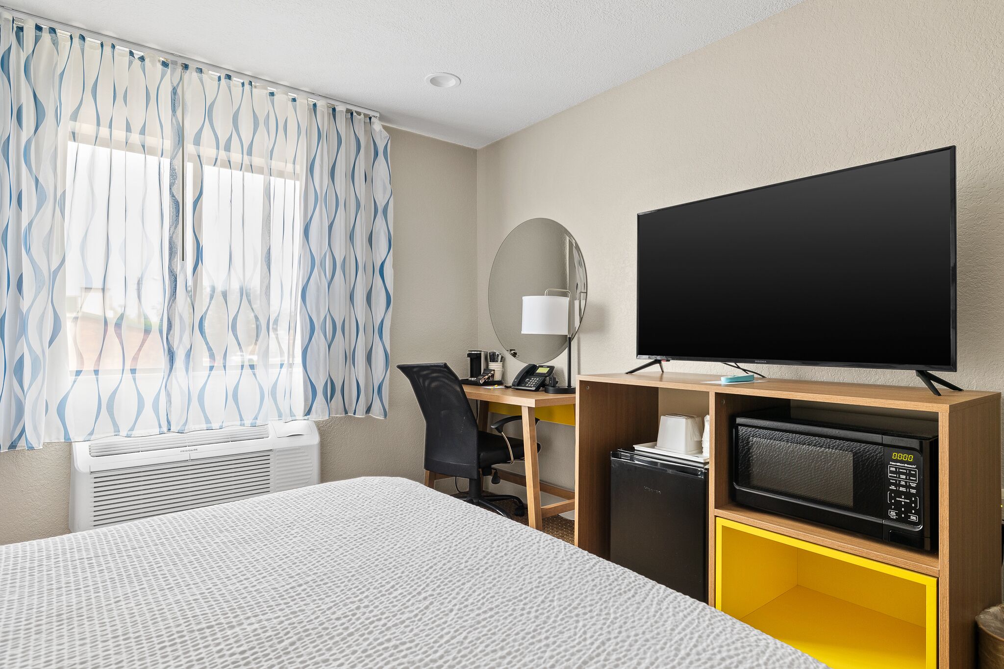 A hotel room with a bed, a desk with a computer, a television, a microwave, and a window with curtains.
