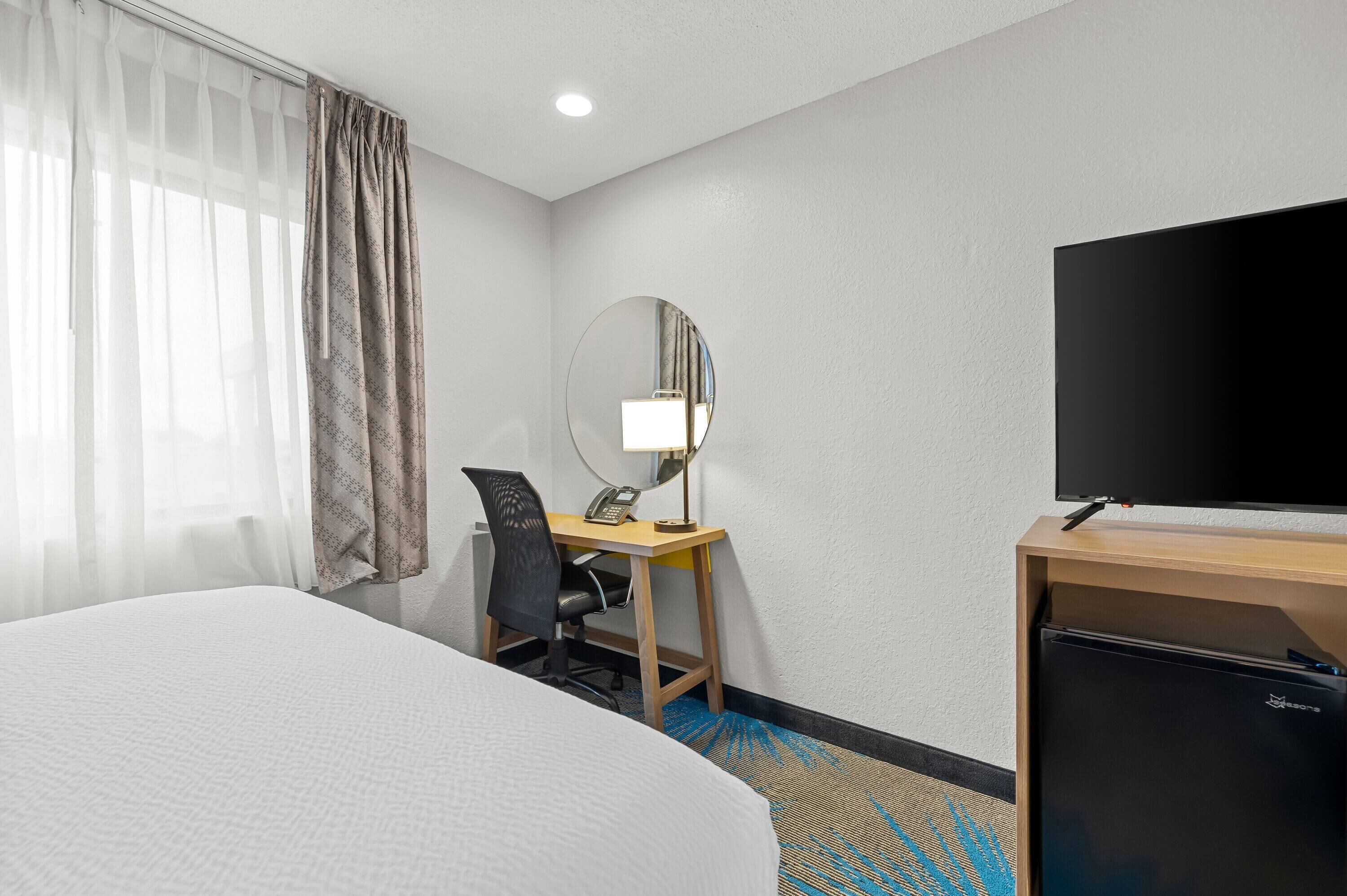 A hotel room with a large bed, a desk with a chair, a mirror, a television, and a refrigerator.