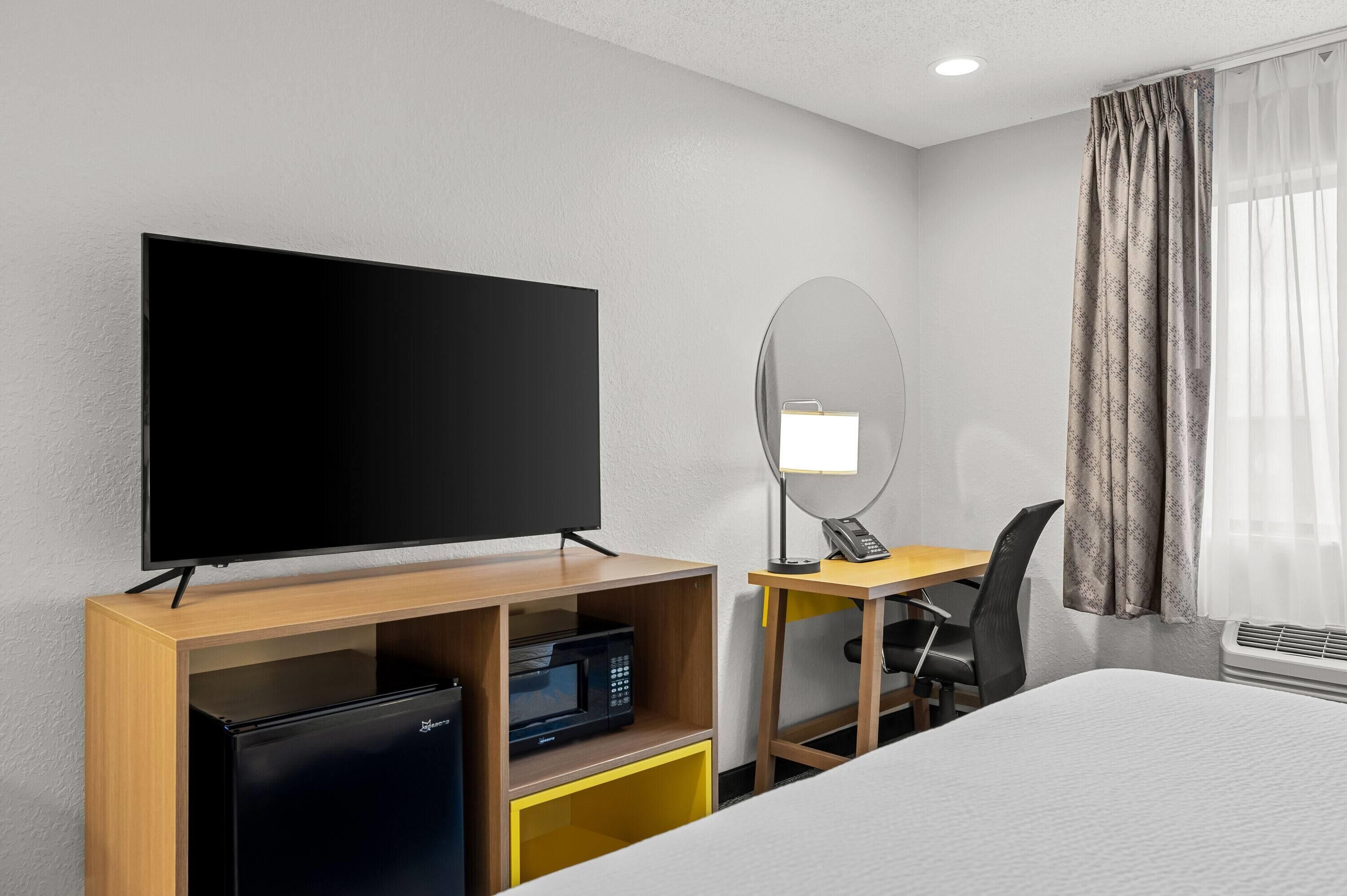 A hotel room with a flat-screen TV, a desk with a chair, a microwave, a mini-fridge, a bed, a mirror, and a window with curtains.