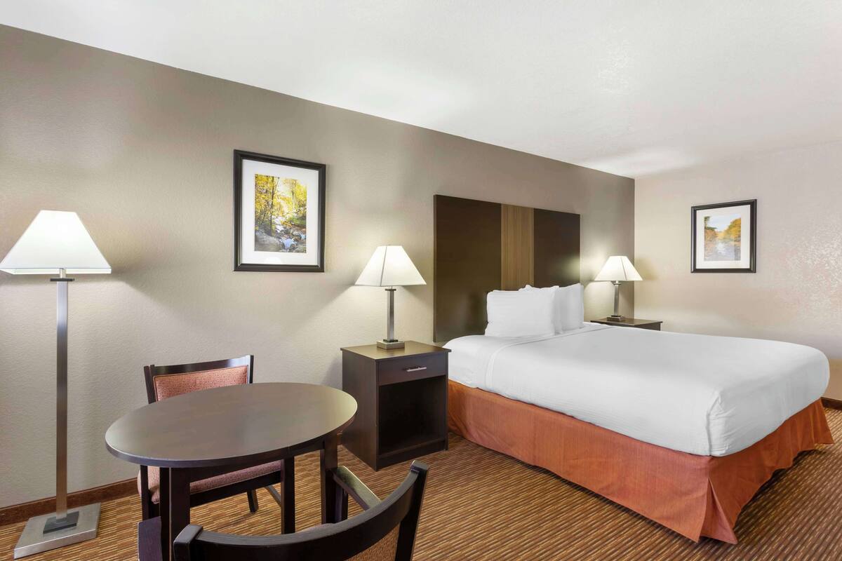 A hotel room with a bed, a desk, two chairs, a lamp, and two framed pictures on the wall.