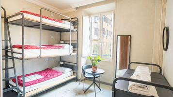 A dormitory room with bunk beds, a small table with a plant, and a window with a view of buildings.