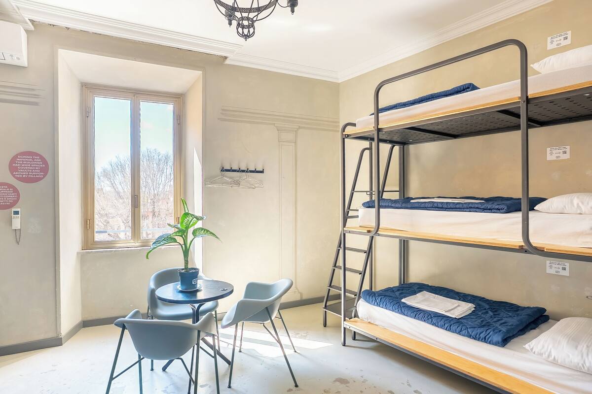 Une chambre de dortoir avec des lits superposés, une petite table avec des chaises, une plante en pot et une fenêtre donnant sur un paysage agréable.