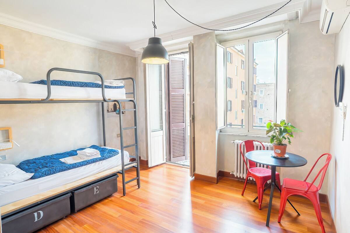 Une petite pièce avec des lits superposés, une petite table avec des chaises, une plante en pot et une vue par la fenêtre.