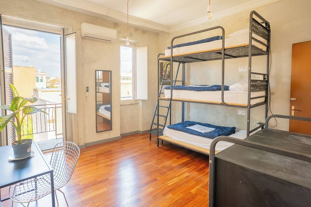 Une chambre avec des lits superposés, une plante en pot, un bureau avec une chaise et une fenêtre donnant sur des immeubles.
