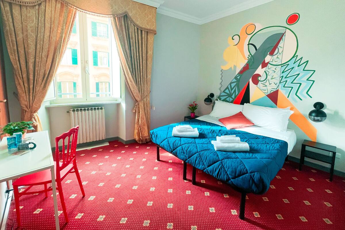 Une pièce comprenant un lit, une moquette rouge, une fenêtre avec des rideaux, un bureau avec une chaise et un mur orné d’une fresque colorée.