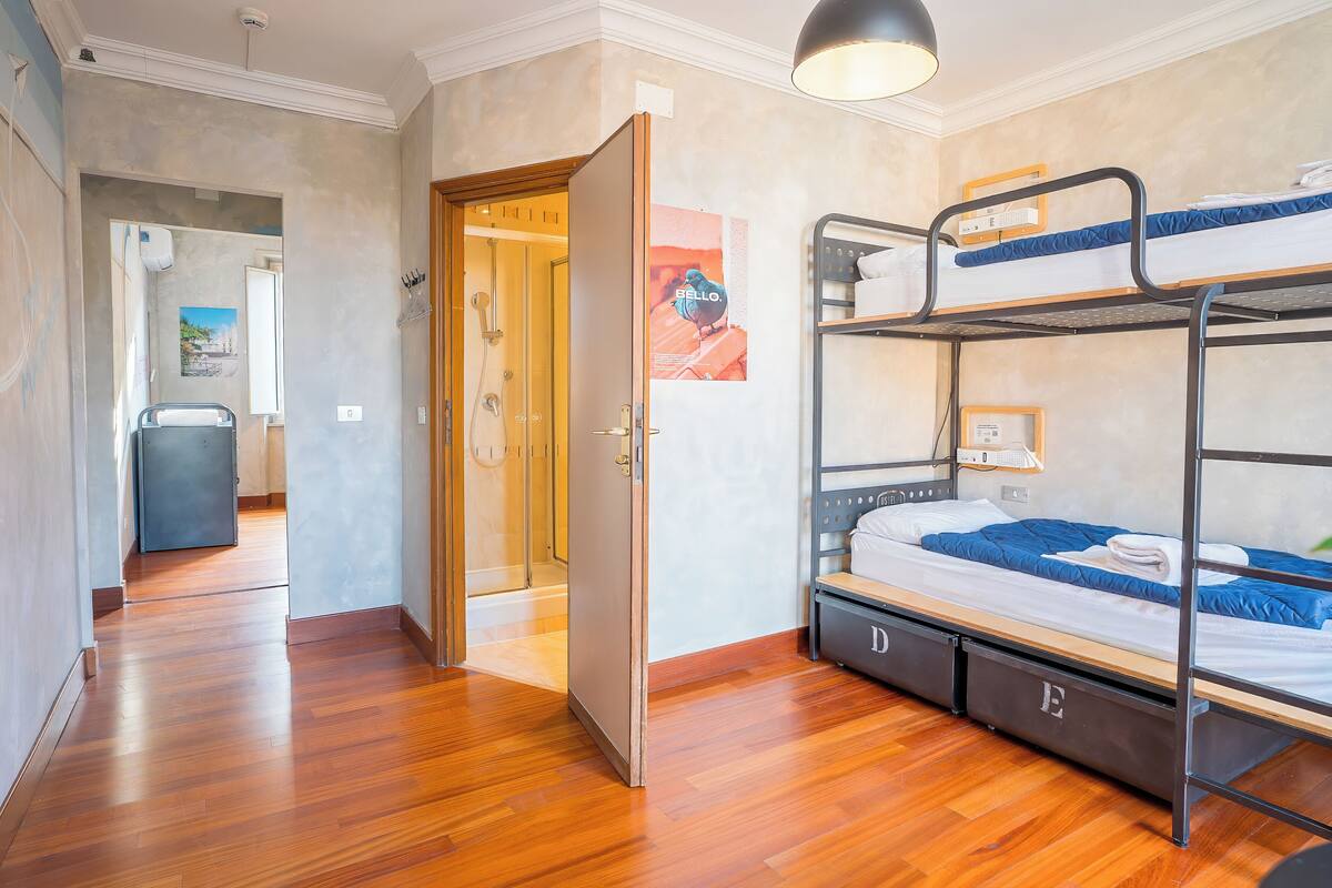 Une pièce avec un lit superposé, un plancher en bois, une porte donnant sur une autre pièce et un espace douche.