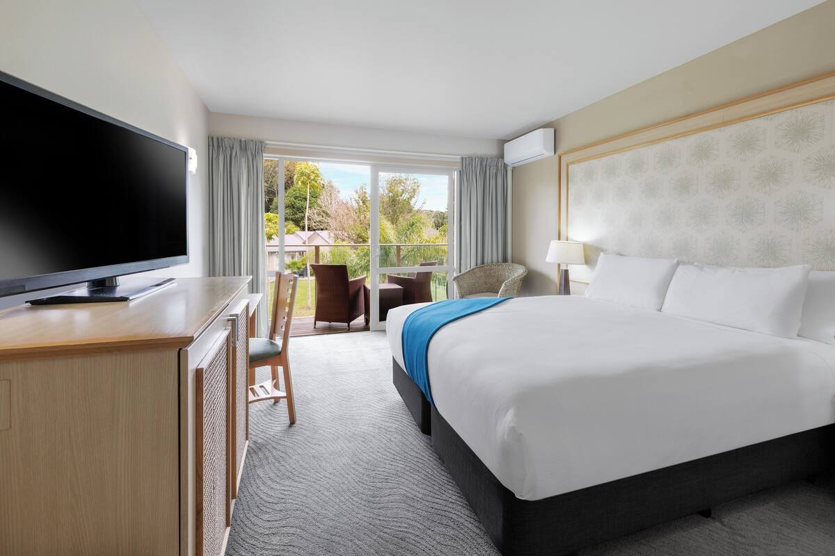 A hotel room with a large bed, a desk with a flat-screen TV, a chair, and a view of the outdoors.