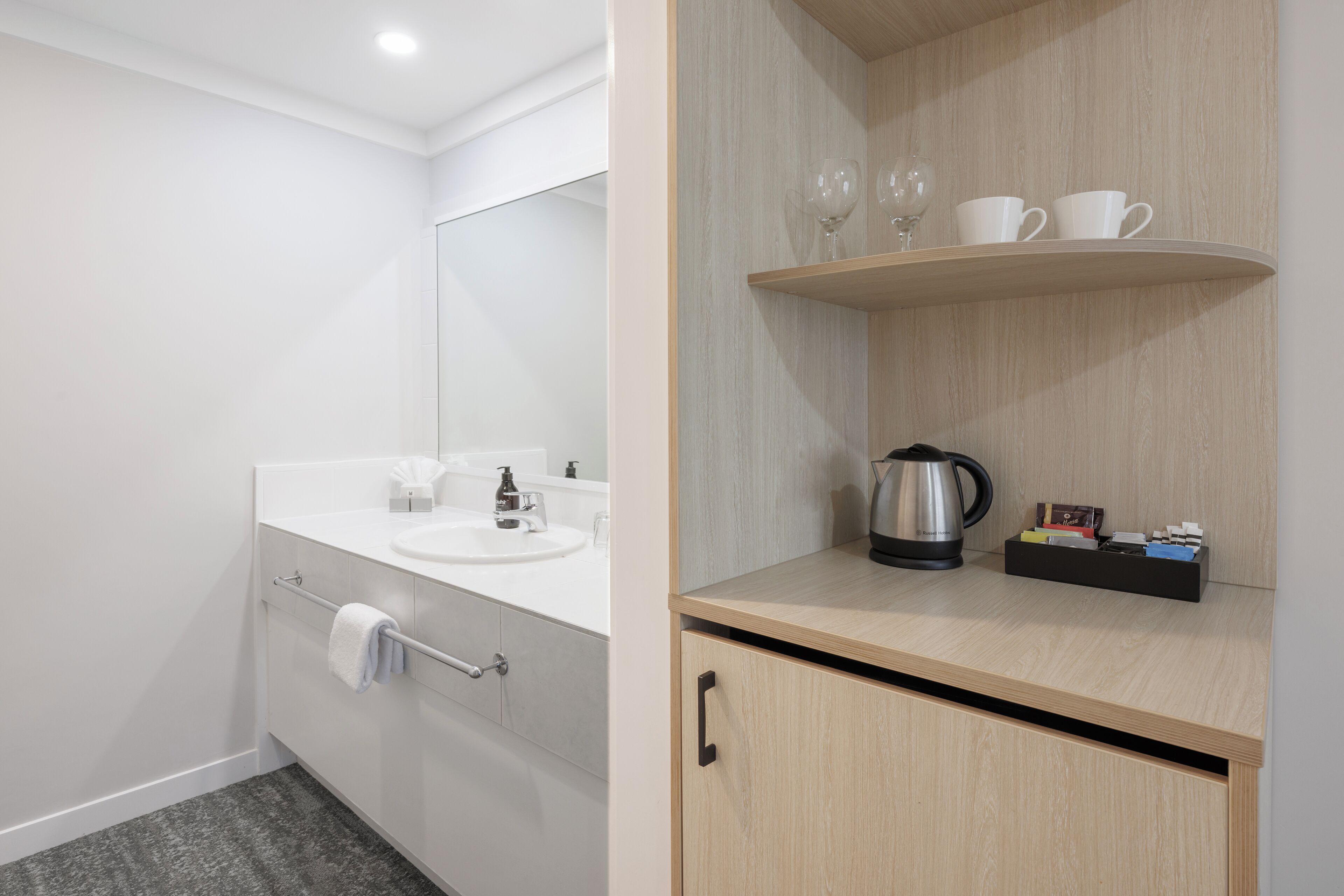 A modern bathroom with a white vanity, a mirror, a towel rack, and a shelf with cups and a kettle.