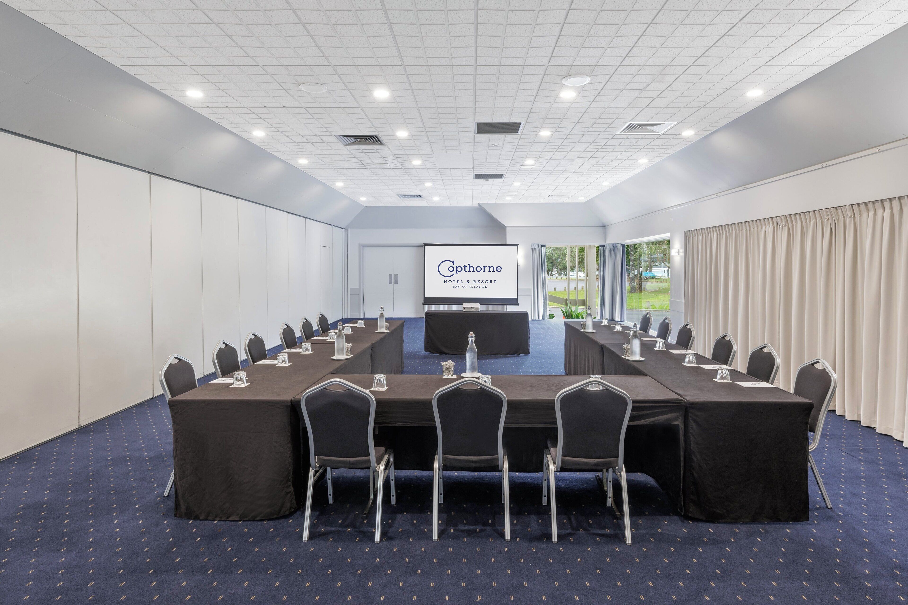 A conference room with a long table, chairs, a podium, and a screen displaying the hotel name.