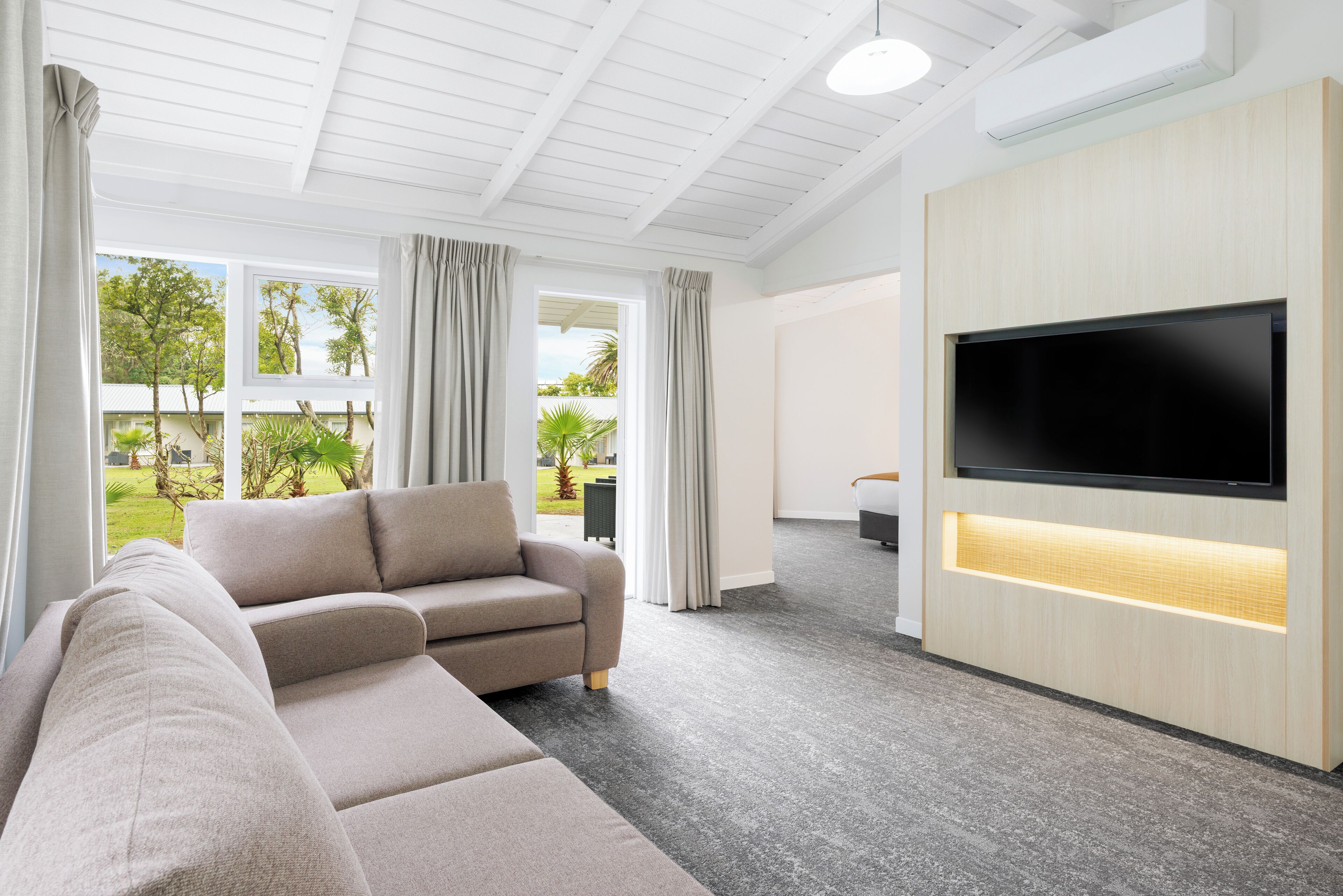 A modern living room with a sectional sofa, a flat-screen TV, and large windows offering a view of greenery.