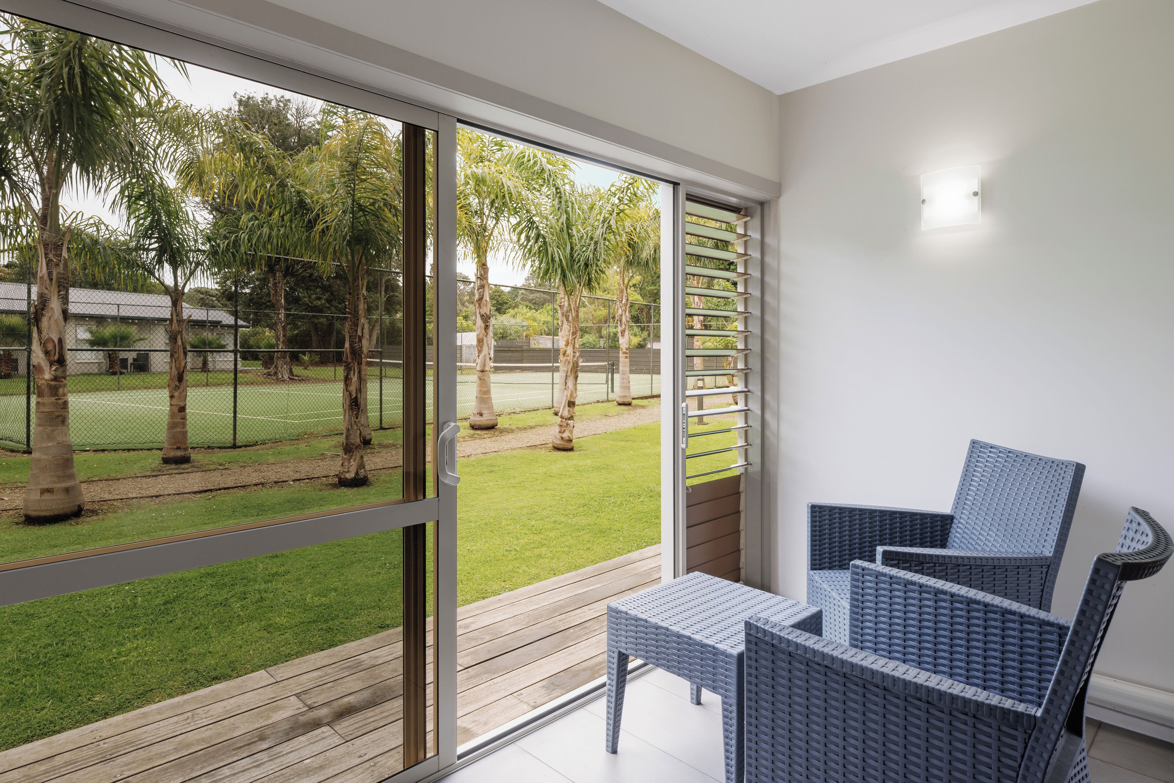 Varanda com mobiliário de vime, vista para uma quadra de tênis e uma parede branca com luminária.