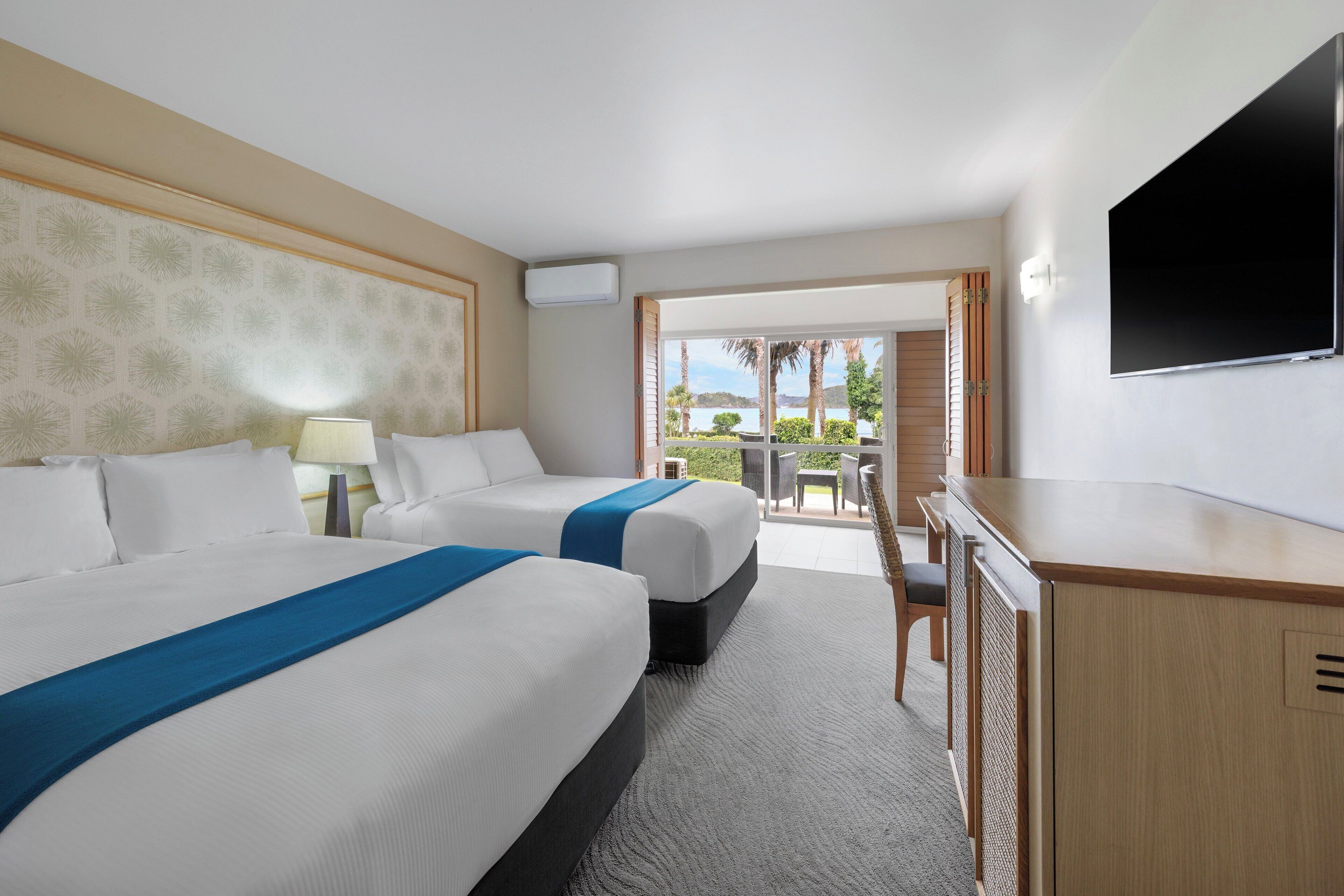 A hotel room with two beds, a desk, a TV, and a balcony with a view of the outdoors.