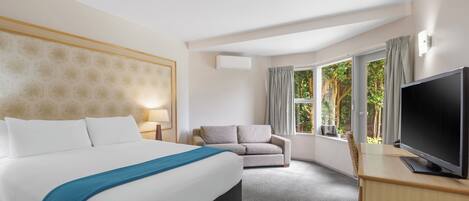 A hotel room with a large bed, a grey armchair, a television, and a window with curtains.