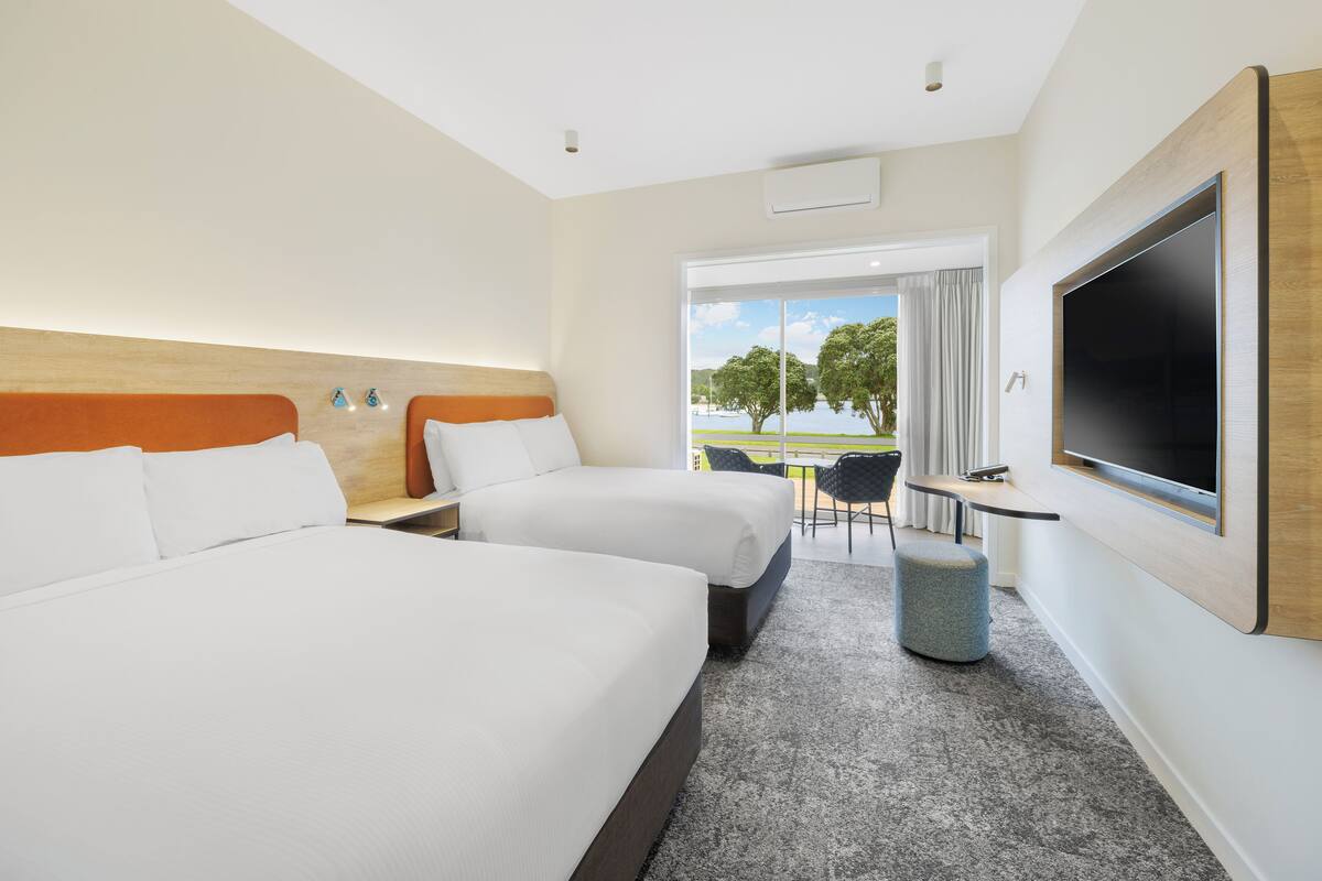 A hotel room with two beds, a TV, a small table, and a view of the outdoors.
