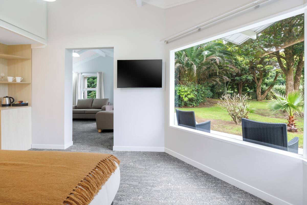 A bedroom with a large window, a bed, a TV, and a view of a garden.