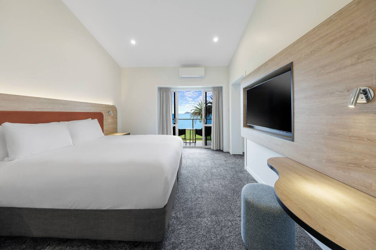 A modern hotel room with a large bed, a flat-screen TV mounted on the wall, and a view of the outdoors through a large window.
