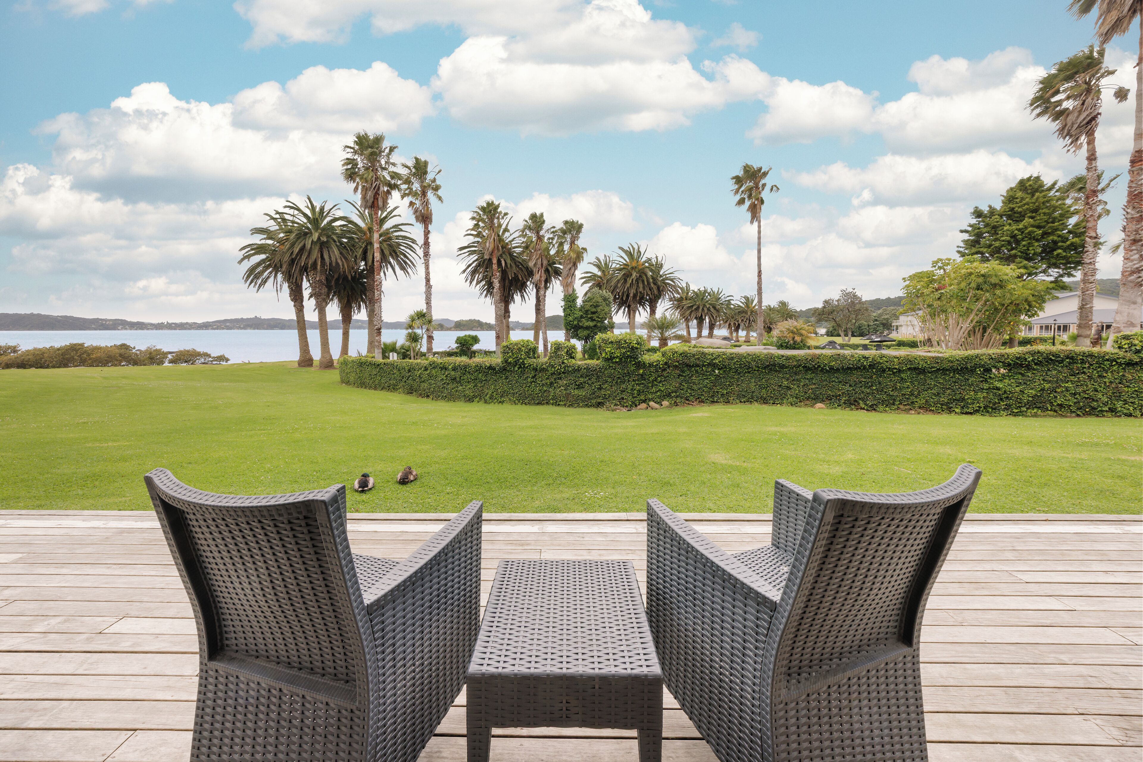 A deck with wicker chairs overlooking a well-manicured lawn and palm trees, with a view of the water and distant hills.