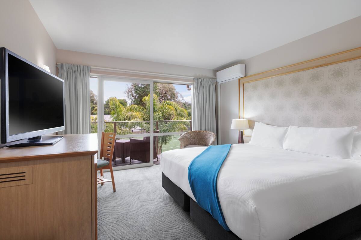 A hotel room with a large bed, a flat-screen TV, a desk, and a balcony with outdoor seating.