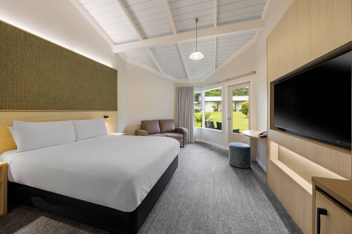 A modern hotel room with a large bed, a flat-screen TV, a seating area with a sofa and armchair, and a view of the outdoors through a large window.
