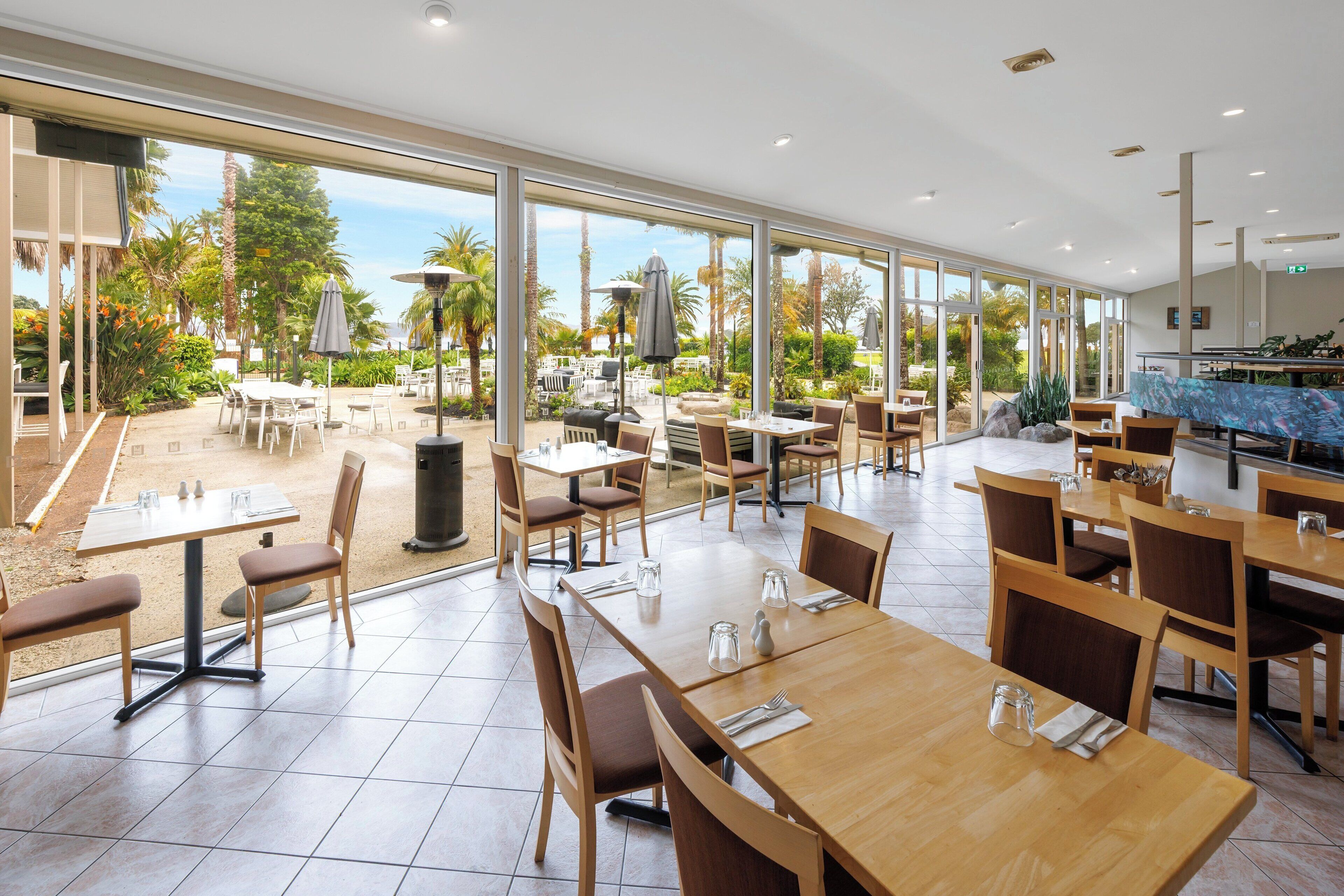 A spacious indoor dining area with large windows, wooden tables, and chairs, overlooking a landscaped outdoor area.