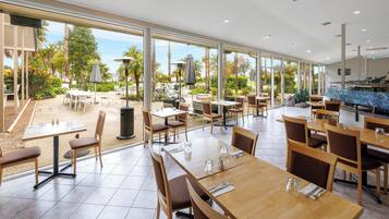 A spacious indoor dining area with large windows, wooden tables, and chairs, overlooking a landscaped outdoor area.