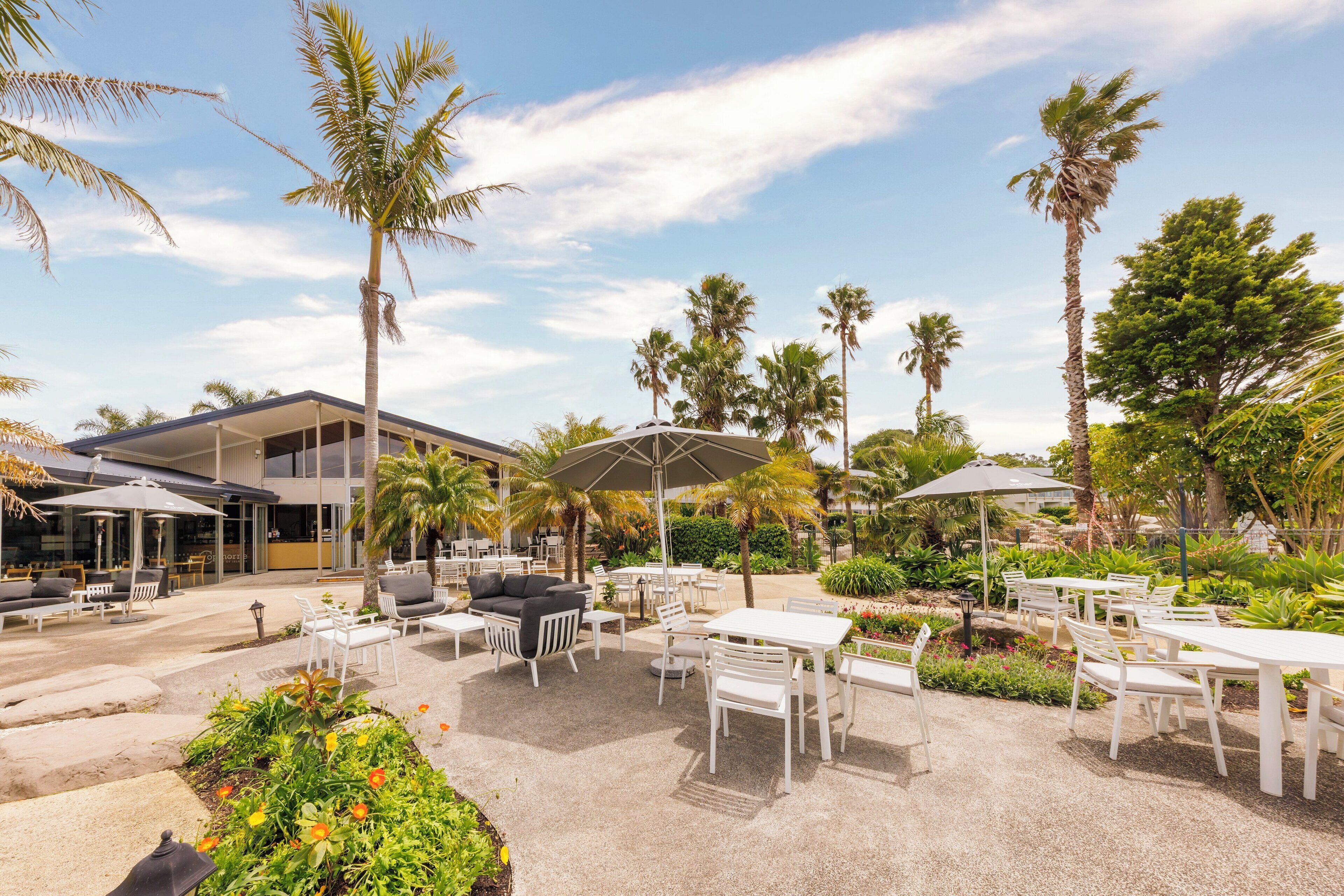 An outdoor patio area with white tables and chairs, palm trees, and a building in the background.