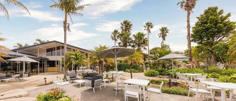 An outdoor patio area with white tables and chairs, palm trees, and a building in the background.