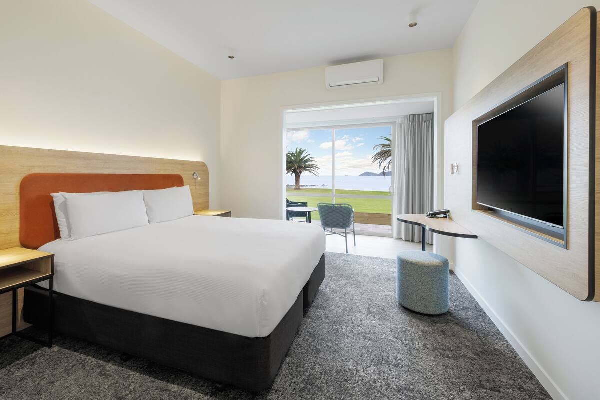 A modern hotel room with a large bed, a flat-screen TV, a small table, and a view of the sea through a large window.