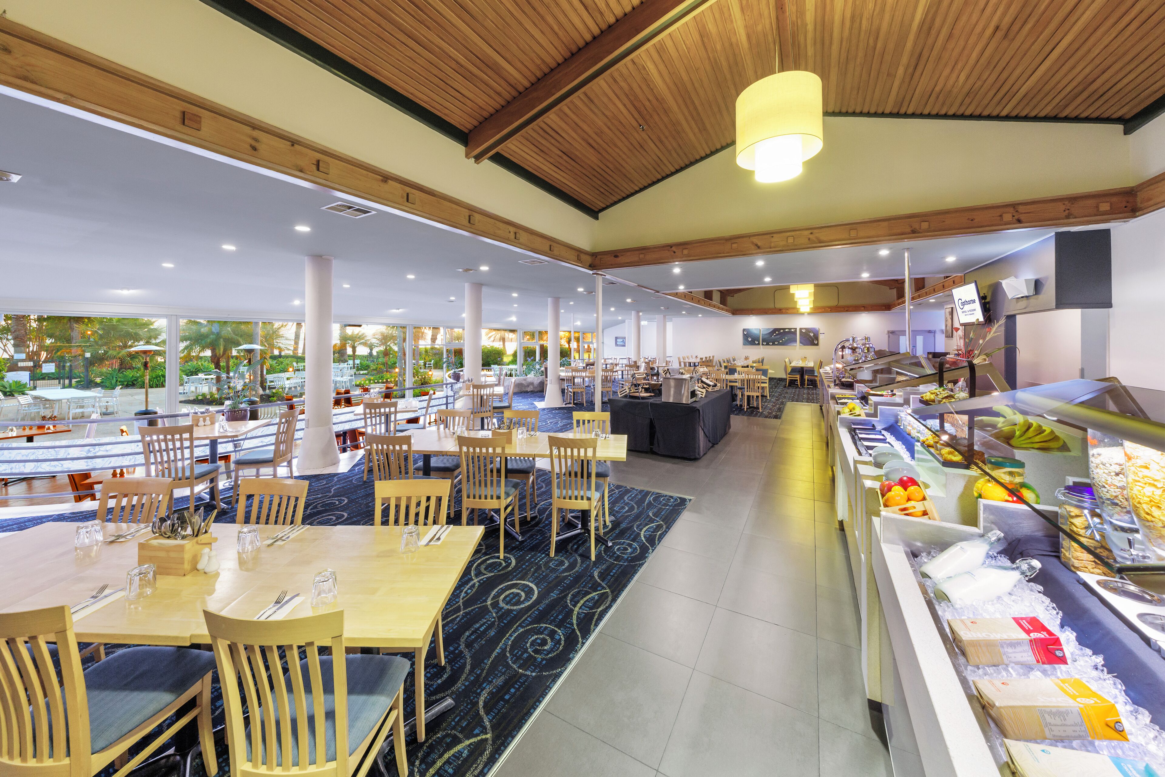Un restaurant spacieux avec des tables et des chaises en bois, un espace buffet proposant une variété de plats, et de grandes fenêtres offrant une vue sur l’extérieur.