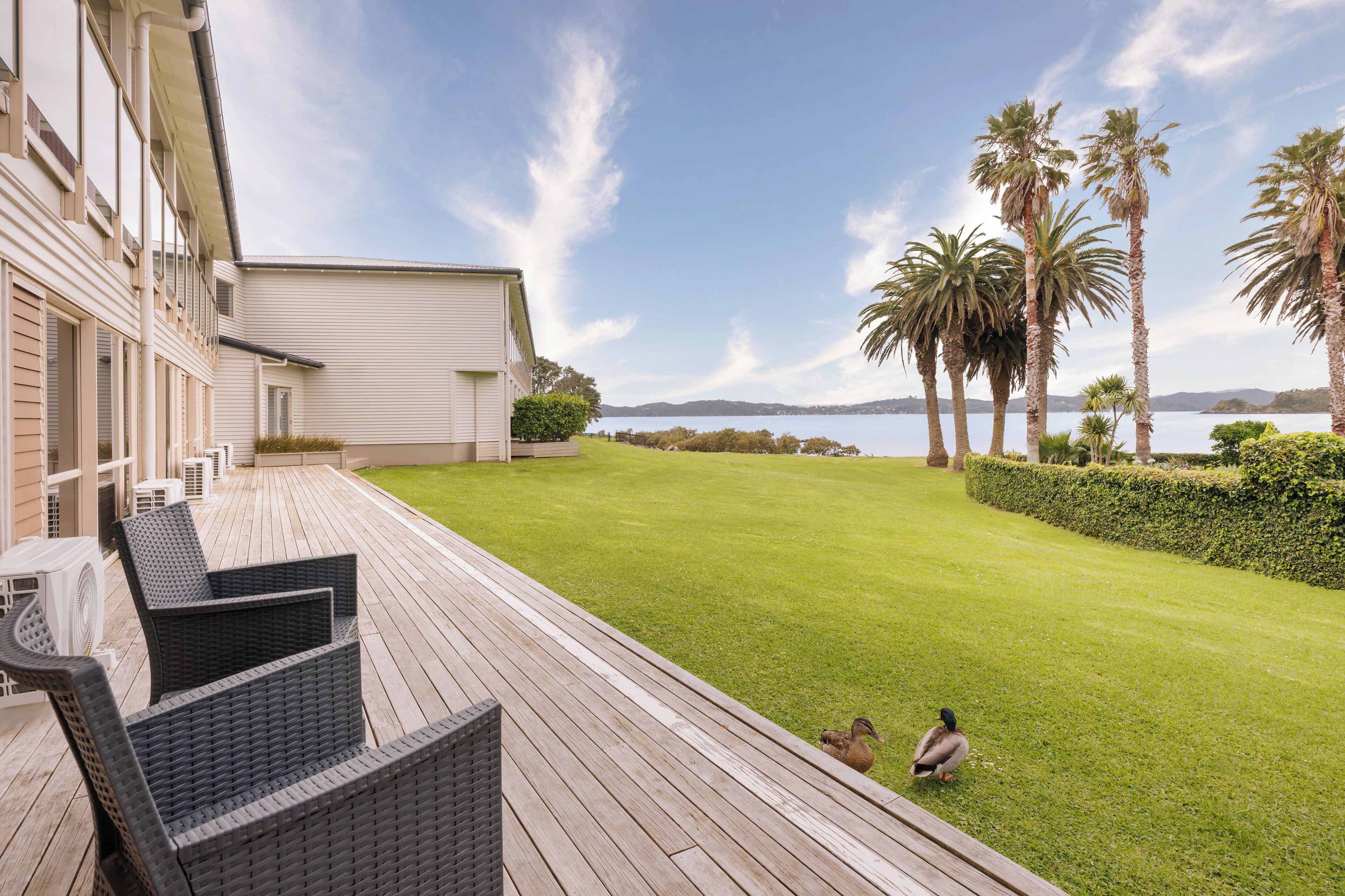 A patio with wicker furniture, a well-maintained lawn, and a view of a body of water with palm trees.