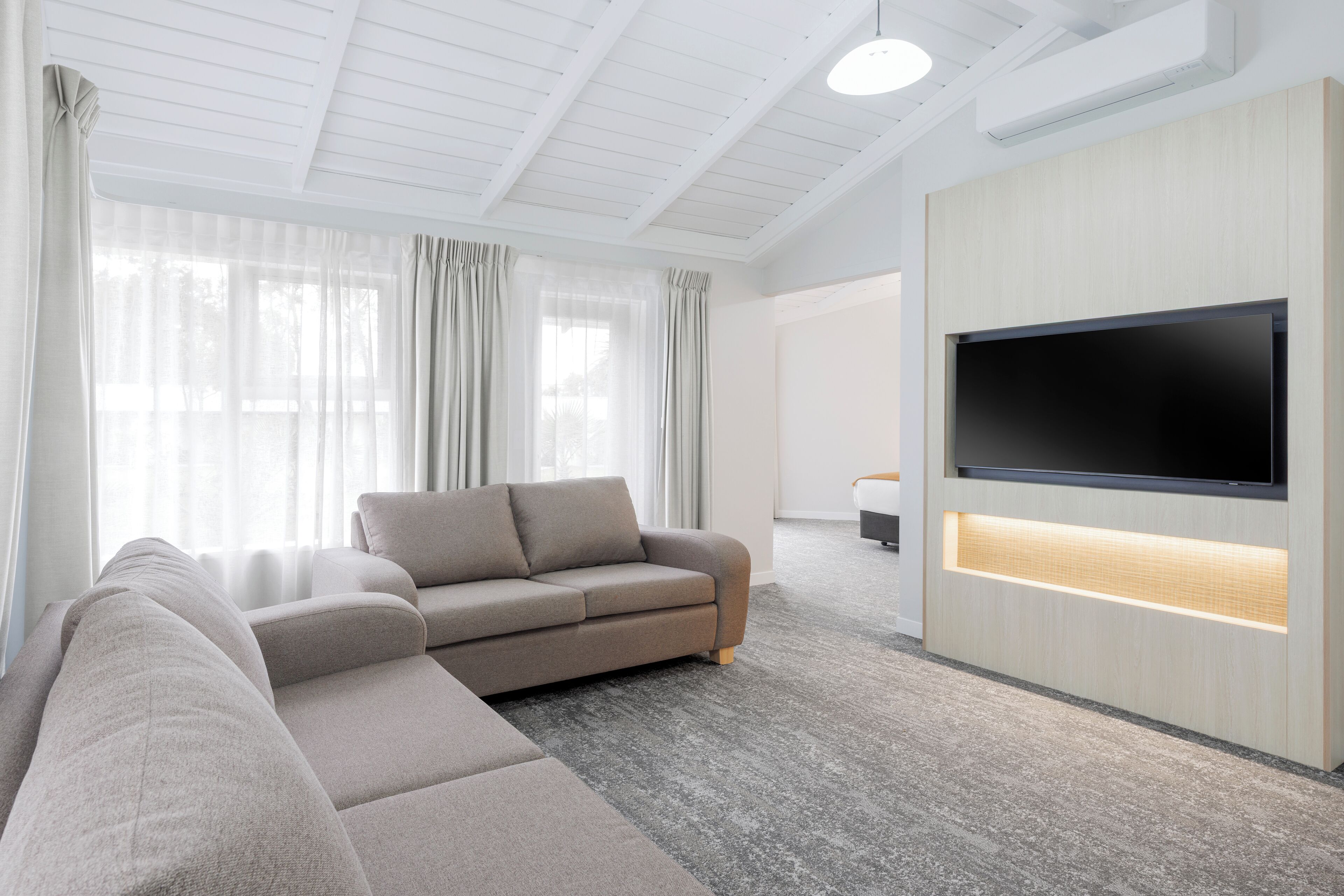 A modern living room with a sectional sofa, a flat-screen TV mounted on a wooden wall, and large windows with sheer curtains.