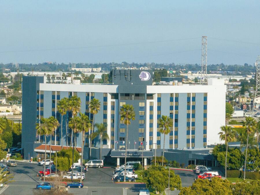 A large, multi-story hotel with a prominent logo on the roof, surrounded by a parking lot and palm trees.