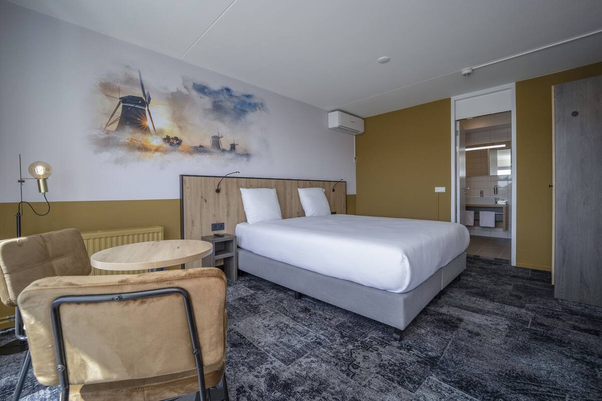 Une chambre d’hôtel avec un lit, une table ronde, une chaise, un radiateur et un mur orné d’un tableau représentant un paysage urbain.