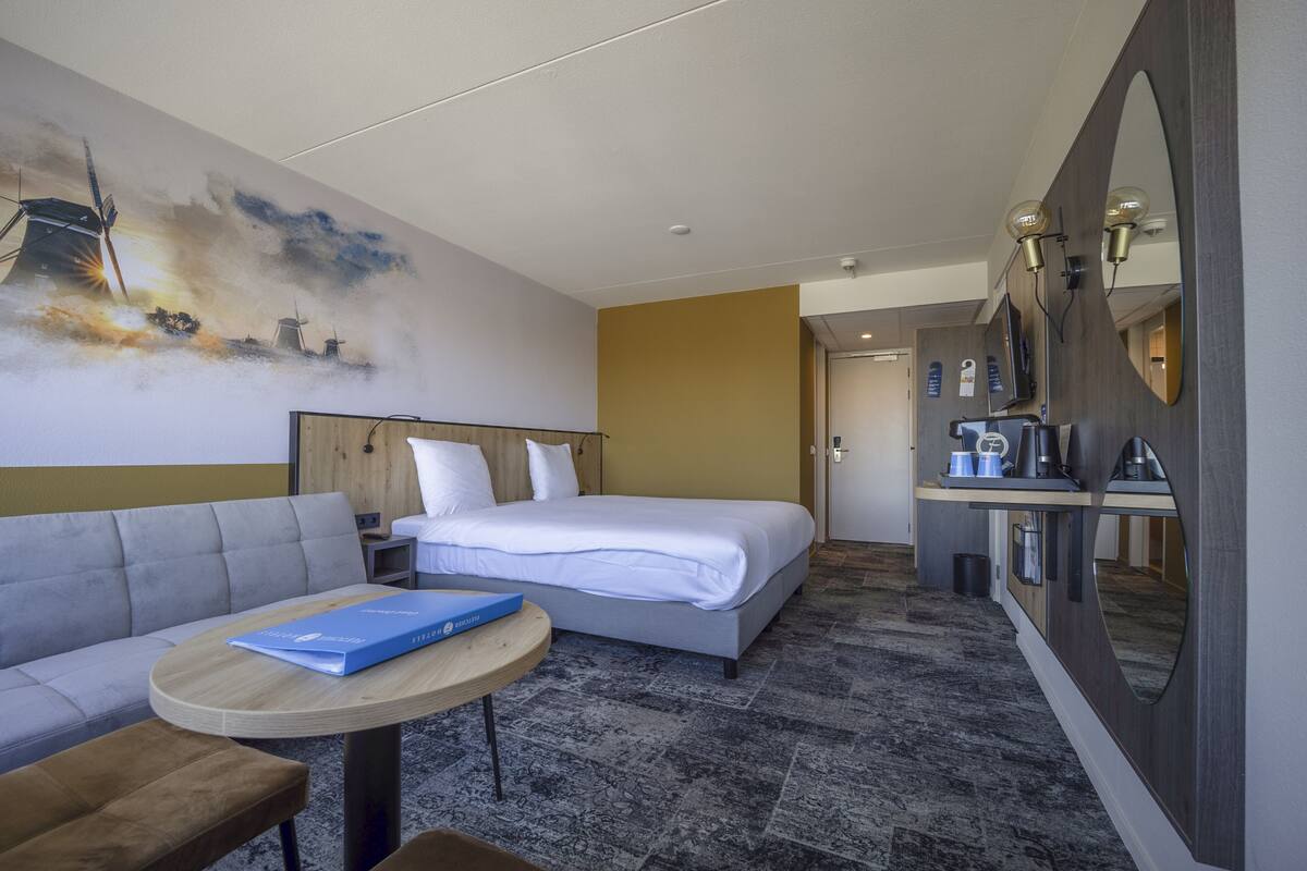 Une chambre d’hôtel avec un lit, un canapé, une petite table avec un livre, un miroir et un mur orné d’un tableau.
