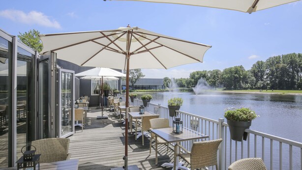 Een terras met buitenmeubilair, een grote parasol en uitzicht op een meer met een fontein.