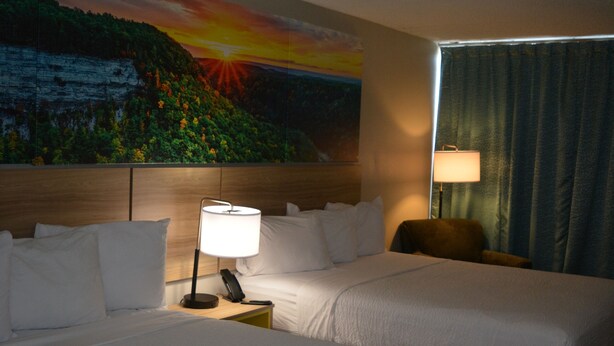 A hotel room with two beds, a nightstand with a lamp, a painting of a sunset on the wall, and a window with curtains. - Days Inn by Wyndham Syracuse (Syracuse)