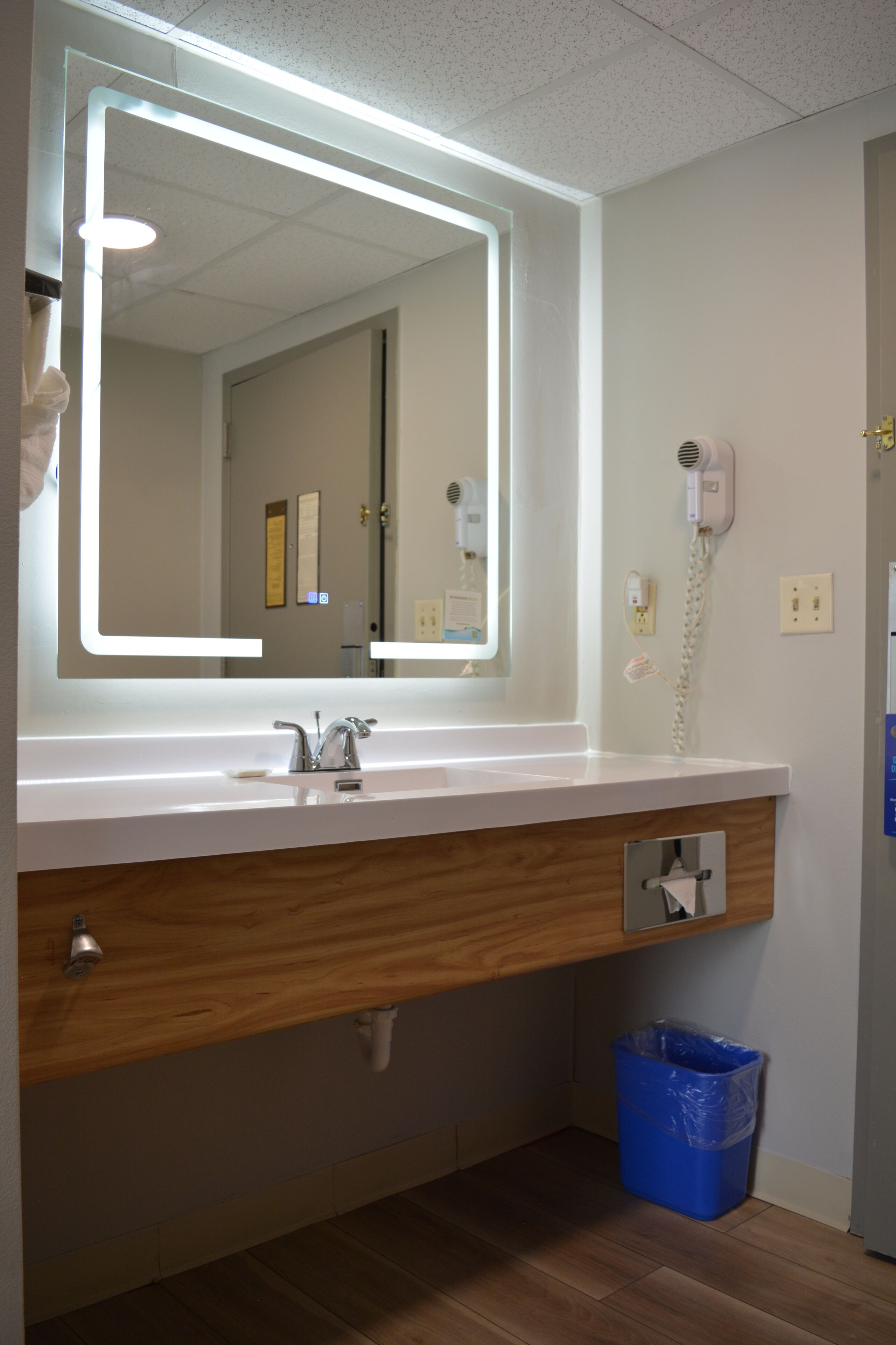 A bathroom with a large mirror, a sink, a hairdryer, and a trash bin.