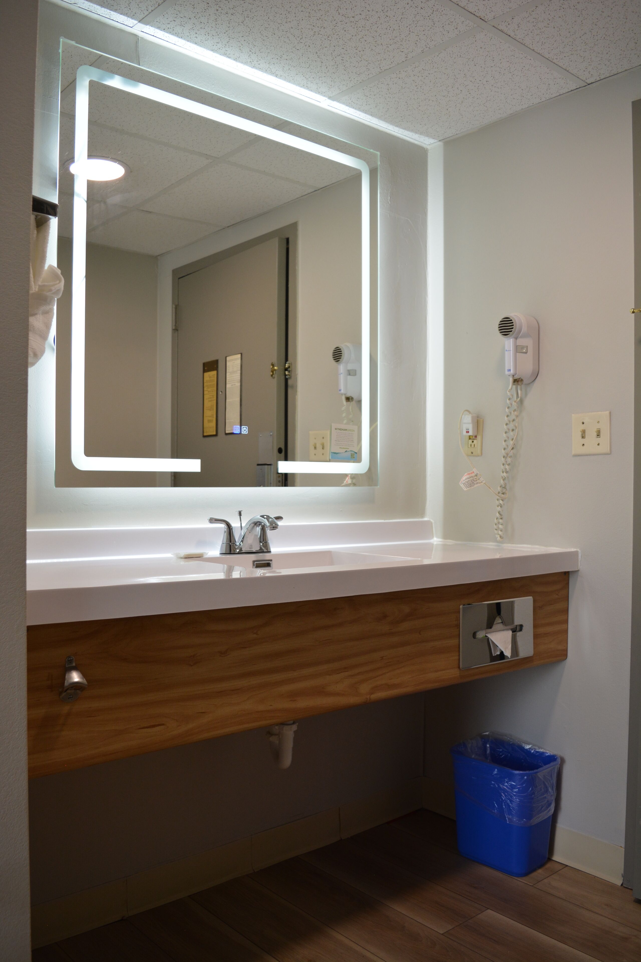 A bathroom with a large mirror, a sink, a hairdryer, and a trash bin.
