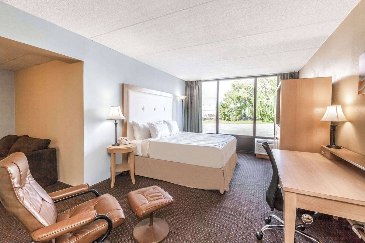 A hotel room with a bed, a desk, a chair, a small table, and a lamp.