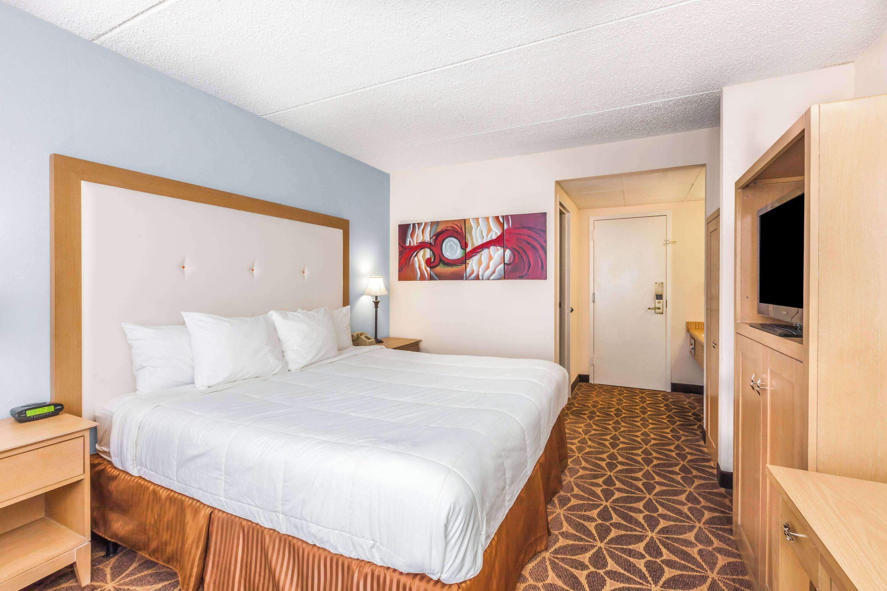 A hotel room with a large bed, a wooden headboard, a flat-screen TV, a desk with a lamp, and a patterned carpet.