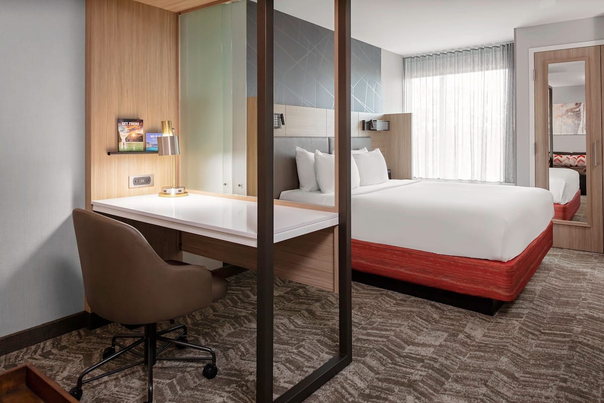 A hotel room with a bed, a desk with a chair, a bedside table, a mirror, and a window with curtains.