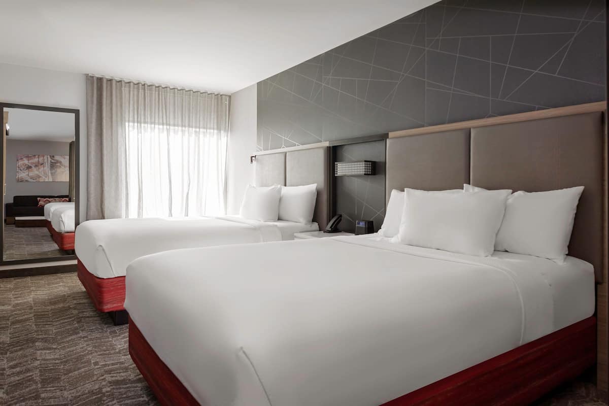 A hotel room with two beds, a large mirror, a bedside table with a lamp, and a view of the cityscape through the window.