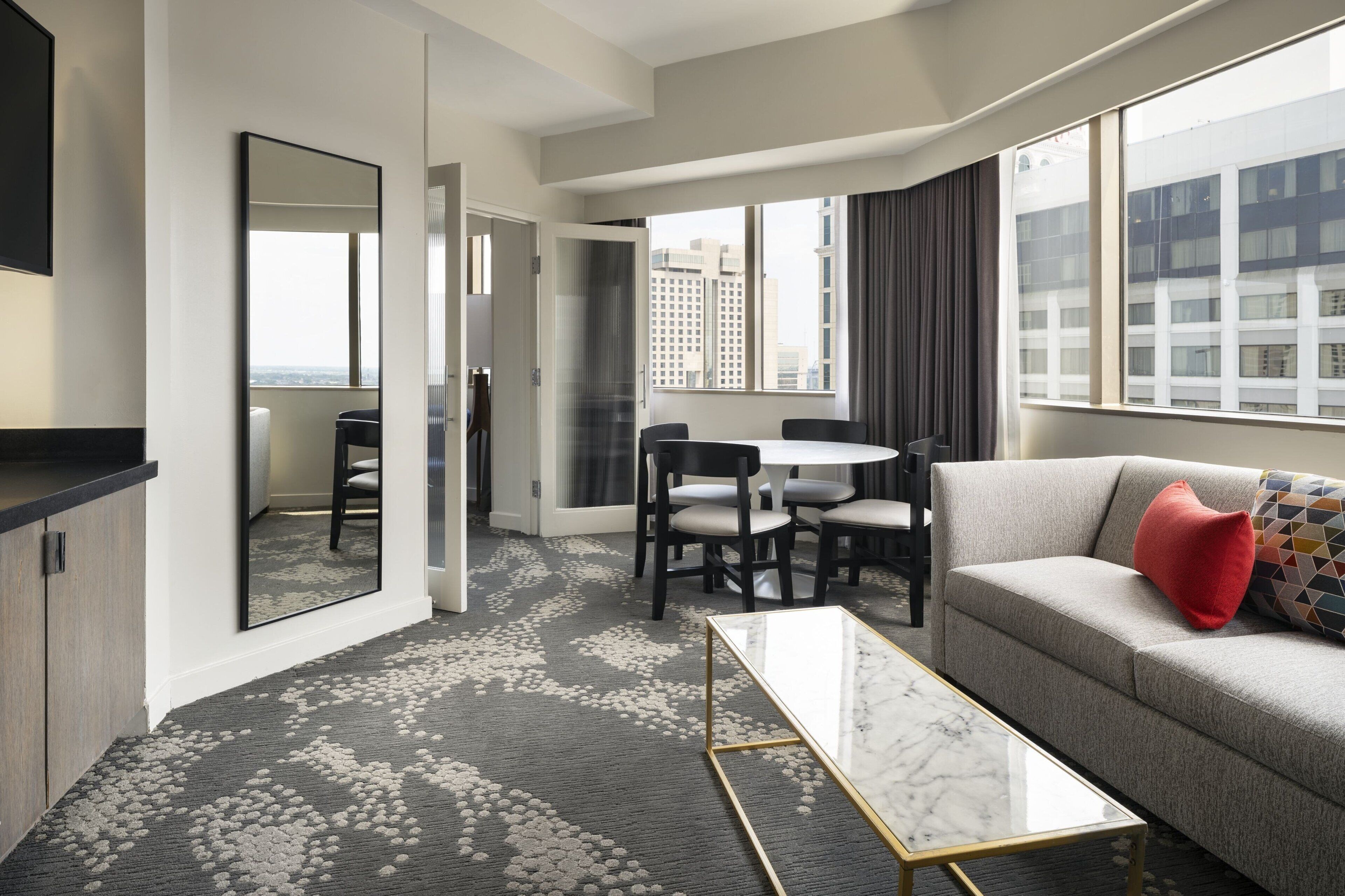 A modern hotel room with a sofa, a coffee table, a dining table with chairs, a large window with curtains, and a city view.