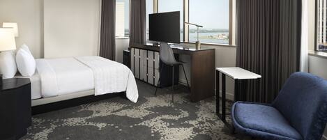A hotel room with a bed, desk, chair, TV, and large windows with a view.