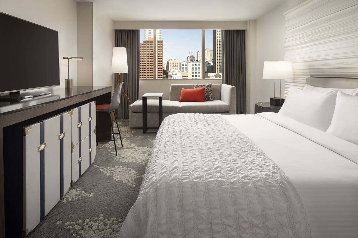 Ein Hotelzimmer mit einem großen Bett, einem Schreibtisch mit Fernseher, einem Sessel, einer Couch und Blick auf die Skyline der Stadt.