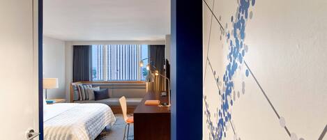 Habitación de hotel con cama, escritorio, silla, lámpara y vistas a la ciudad a través de amplios ventanales.