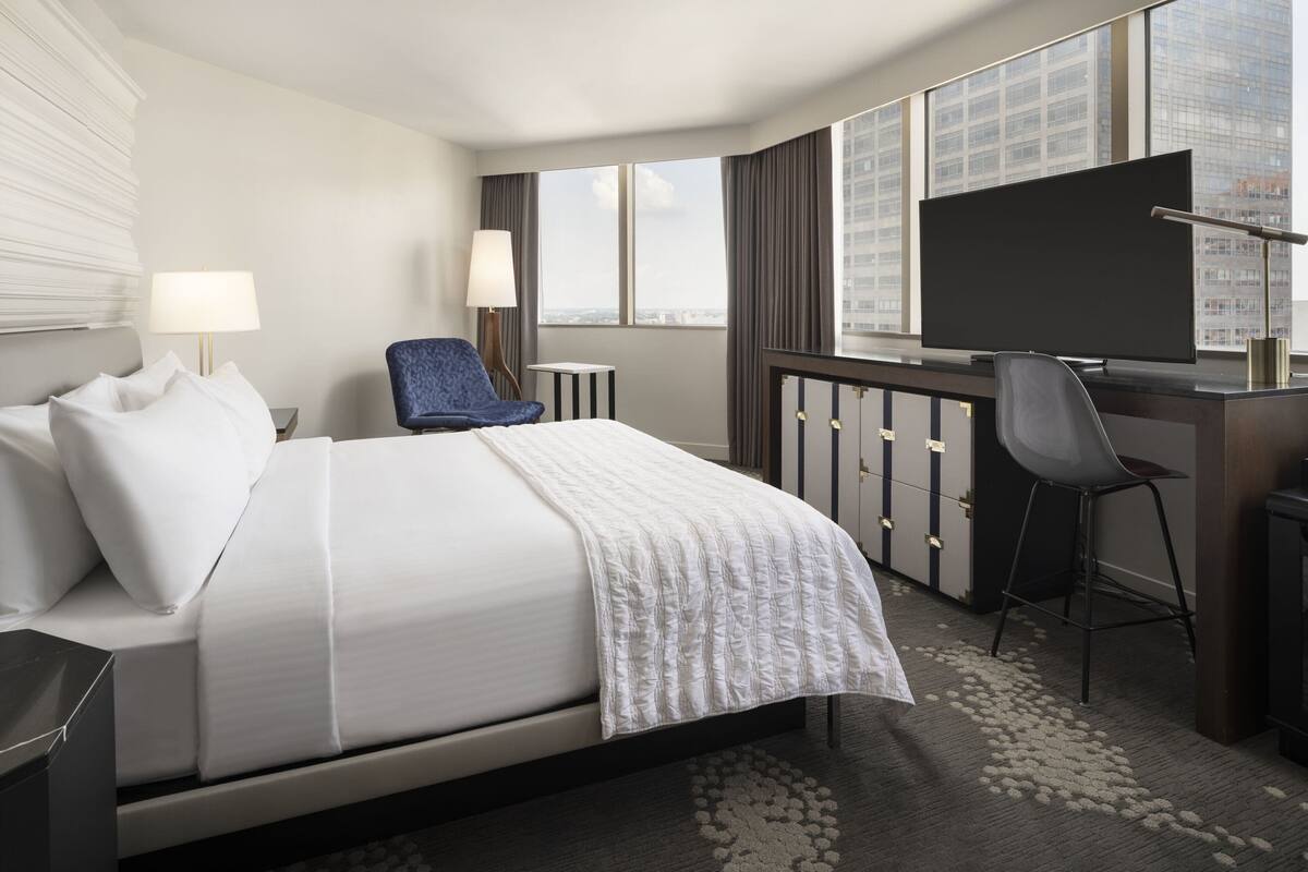 Ein Hotelzimmer mit Bett, Schreibtisch mit Fernseher, Stuhl und großen Fenstern mit Blick auf die Stadt.